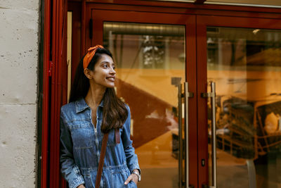 Smiling young woman looking away while standing on window