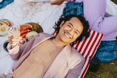 High angle portrait of smiling gay man holding wineglass while lying down in back yard