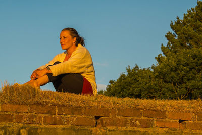 Woman looking away while sitting on brick wall against clear blue sky