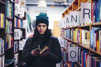 Young woman reading book amidst shelves in library