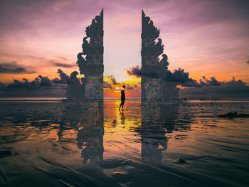 Silhouette of a bali gate on the beach and a person in the middle against the sky at sunset