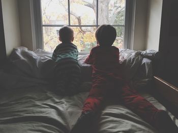 Rear view of children looking through window while lying on bed at home
