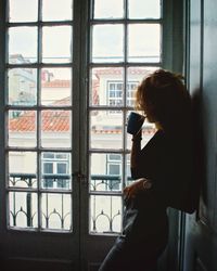 Side view of a woman standing by a window while drinking a cup of tea