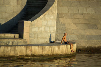 Full length of shirtless man sitting on retaining wall over river