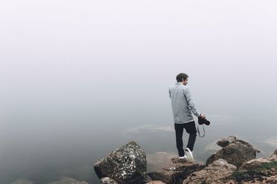 Rear view of mid adult man holding camera while standing on rock during foggy weather