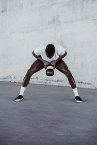 Male athlete lifting kettle bell while standing against white wall