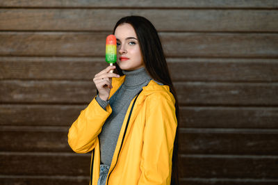 A girl in a yellow raincoat and a gray sweater holds a multicolored ice cream in her hand