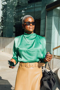 Young ethnic female entrepreneur in trendy clothes standing near stairs of modern business center and looking away