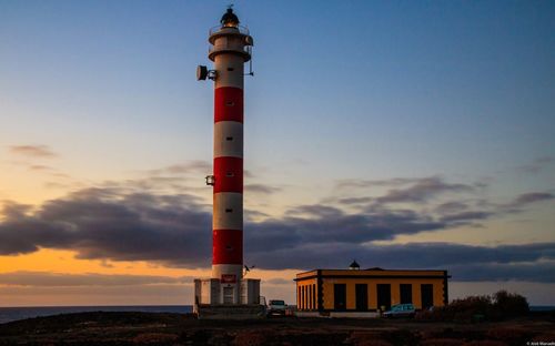 Lighthouse by building against sky during sunset