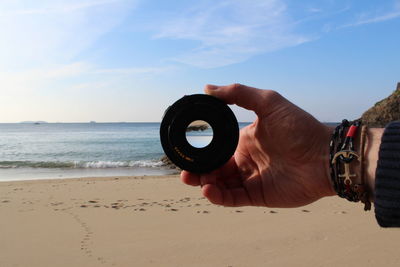 Close-up of hand holding camera lens at beach against sky