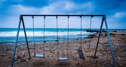 Abandoned swings at sea shore against cloudy sky