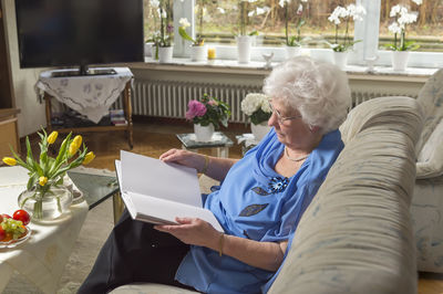 Senior woman reading book while sitting on sofa at home