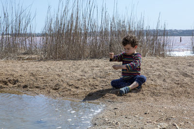 Small boy of 4-5 years playing with sand at the edge of pond on sunny day with colored striped shirt