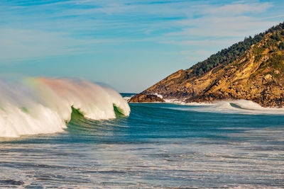 Large rainbow wave in sea against sky with rocky headland in background.