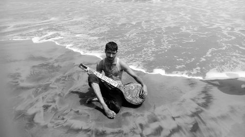 High angle portrait of young man playing sitar while sitting at beach