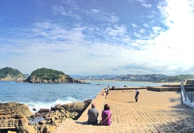 People sitting on promenade at beach against sky