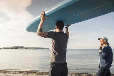 Man carrying paddleboard on head while male friend standing at beach