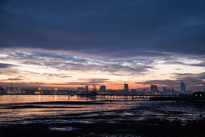 Illuminated cityscape by sea against sky during sunset