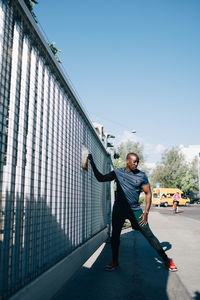 Full length of male athlete stretching by railing on bridge in city during sunny day