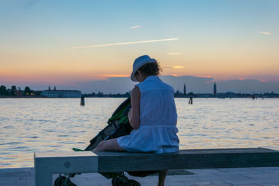 Rear view of woman sitting on bench against sky during sunset