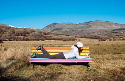 Full body side view of female traveler in warm activewear lying on colorful bench and reading book on grassy terrain with mountains and blue sky in background
