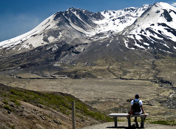 Rear view of man sitting on bench against snowcapped mountain