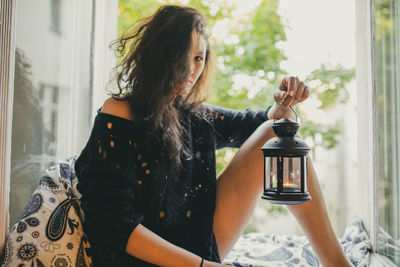Portrait of woman holding lantern while sitting on window sill at home