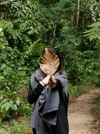 Woman holding dry leaf while standing on footpath amidst trees in forest