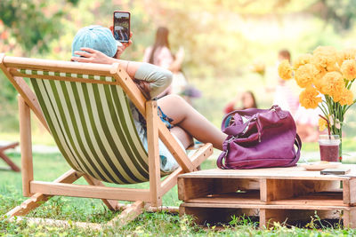Rear view of woman taking selfie through smartphone while sitting on deck chair by table at park