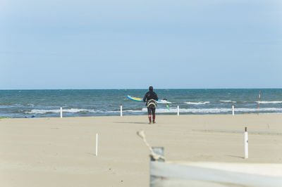 Rear view of person with surfboard walking at beach towards sea against clear sky