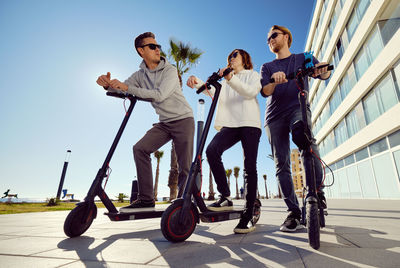 Low angle view of friends using push scooters in city on sunny day