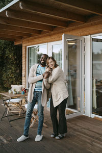 Portrait of happy multiracial couple standing on porch outside house