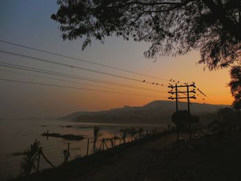 Silhouette electricity pylon by tree against sky during sunset