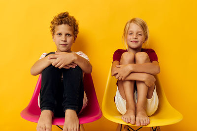 Portrait of sibling sitting on chair against colored background