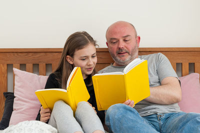 Young school age girl sitting together with her father and discussing yellow textbook. 