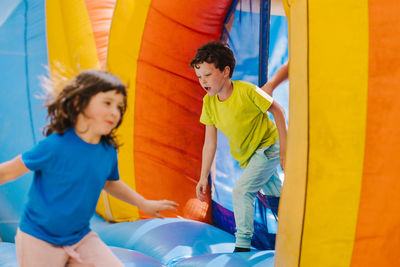 Adorable content little sister and brother with dark hair in t shirts running inside of colorful inflatable trampoline while spending time together on playground on sunny day