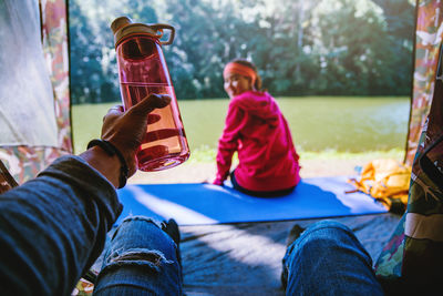 Midsection of man holding bottle with woman sitting in background