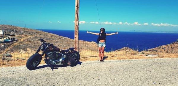 Full length of woman standing by motorcycle on roadside against blue sky