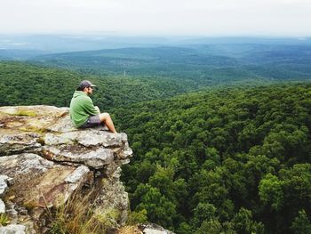 Man sitting on cliff in forest against sky