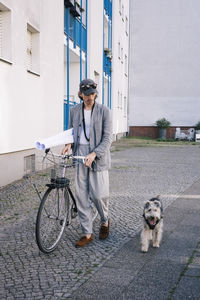 Full length of male architect holding blueprint rolls walking by bicycle and dog on footpath