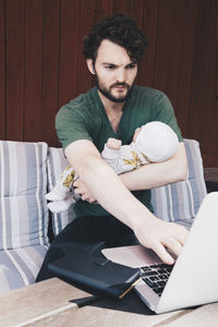 Father with baby boy using laptop while sitting on chair against wall