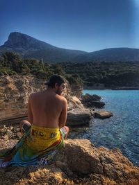 Rear view of shirtless man sitting on rock by sea against sky