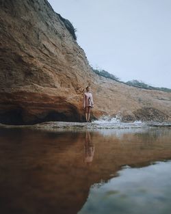 Young woman standing at beach by rock formation against sky