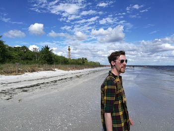 Side view of hipster young man standing on shore at beach against sky