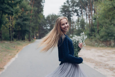 Portrait of young woman holding bouquet while standing on road amidst trees