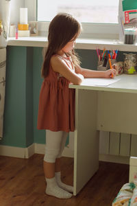 Little serious girl child draws on table standing by window in nursery, elementary home school study