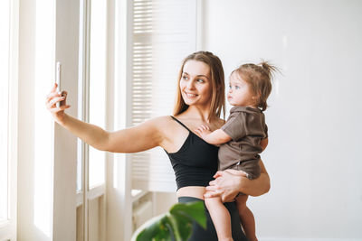 Young mother in sportwear with baby girl on hands taking selfie on mobile phone in bright room