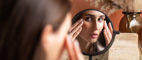 Mirror reflection of woman applying under-eye patch at dressing table closeup