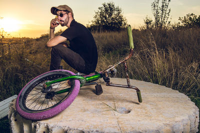 Man sitting with bicycle on field against sky during sunset