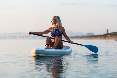 Side view of fit female surfer sitting on paddleboard and rowing against spectacular sun in sunset sky